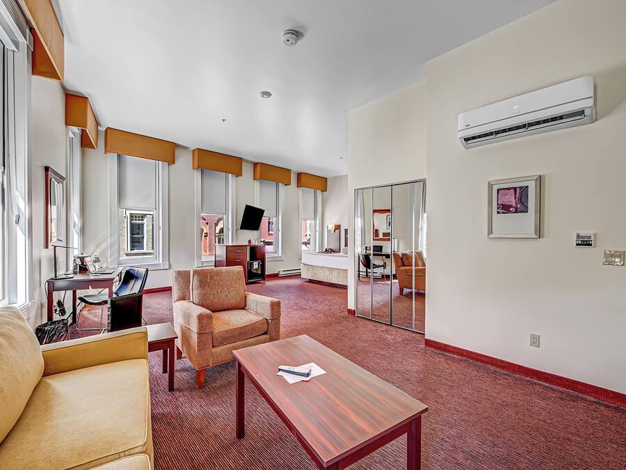 A spacious hotel room with a large window, a flat-screen TV, a sofa, a chair, a small table, and a red carpet.
