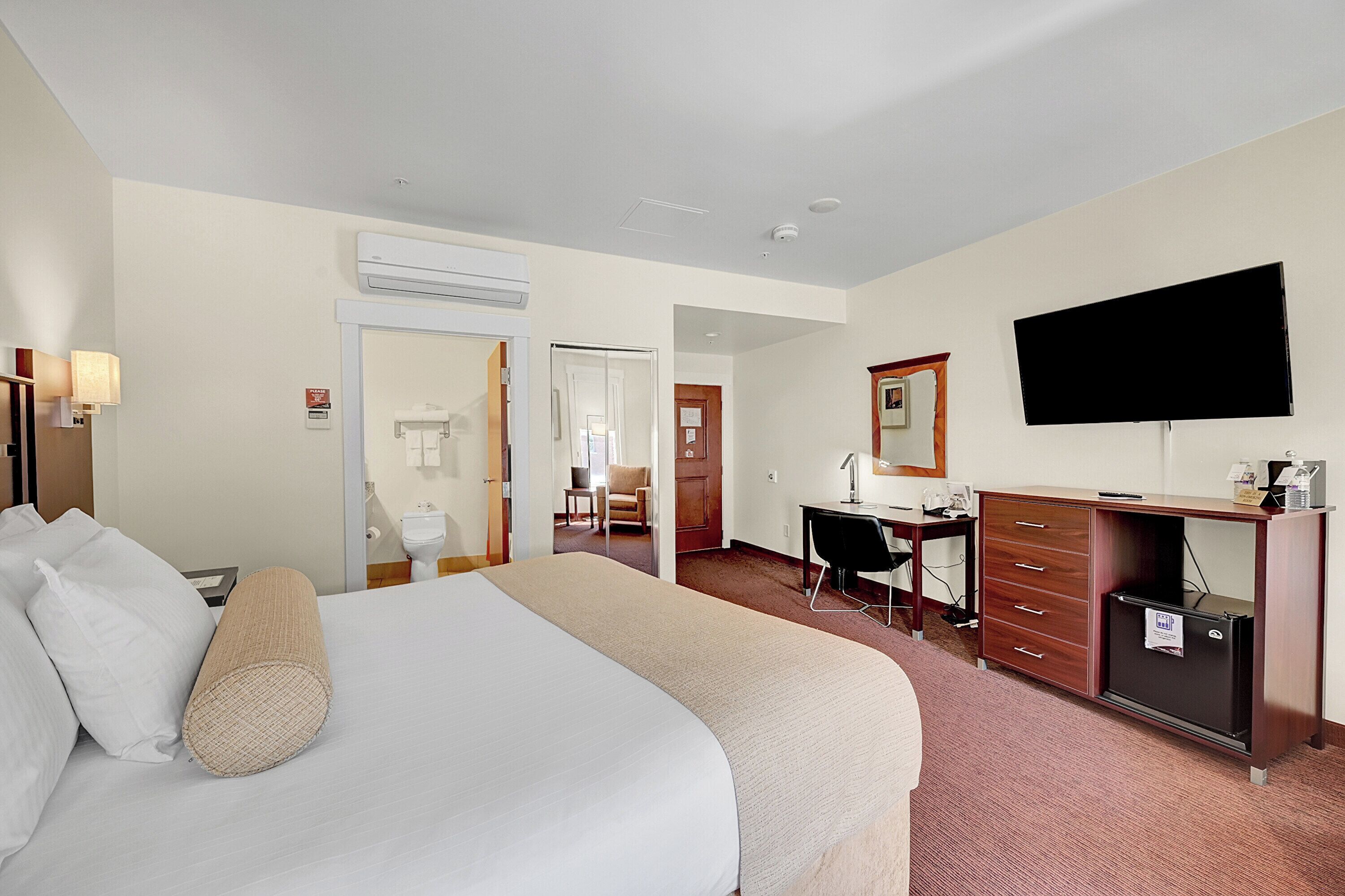 A hotel room with a bed, a desk, a chair, a television, and an air conditioning unit.