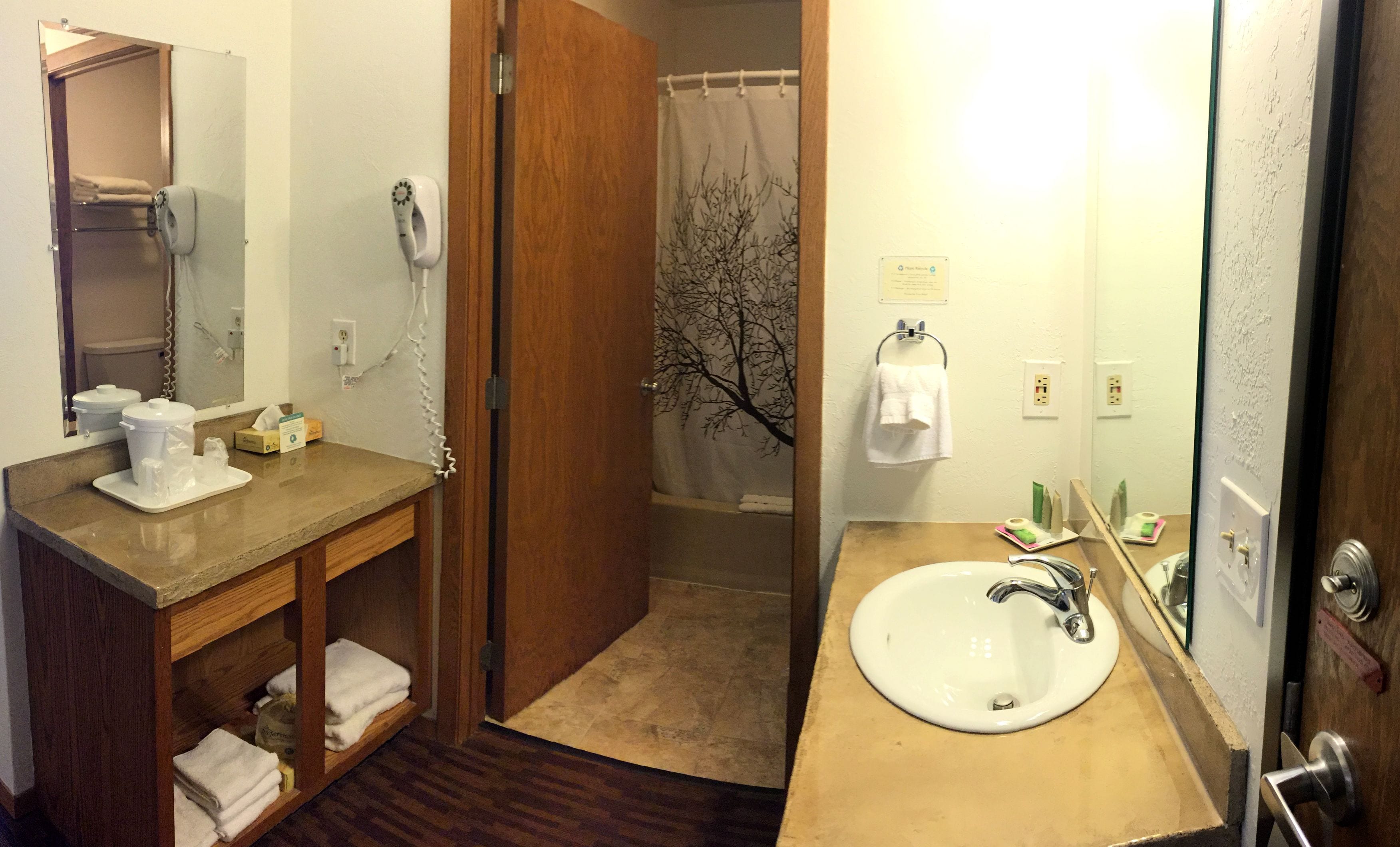 A hotel bathroom with a wooden vanity, a sink, a mirror, a shower curtain with a tree design, and a towel rack.