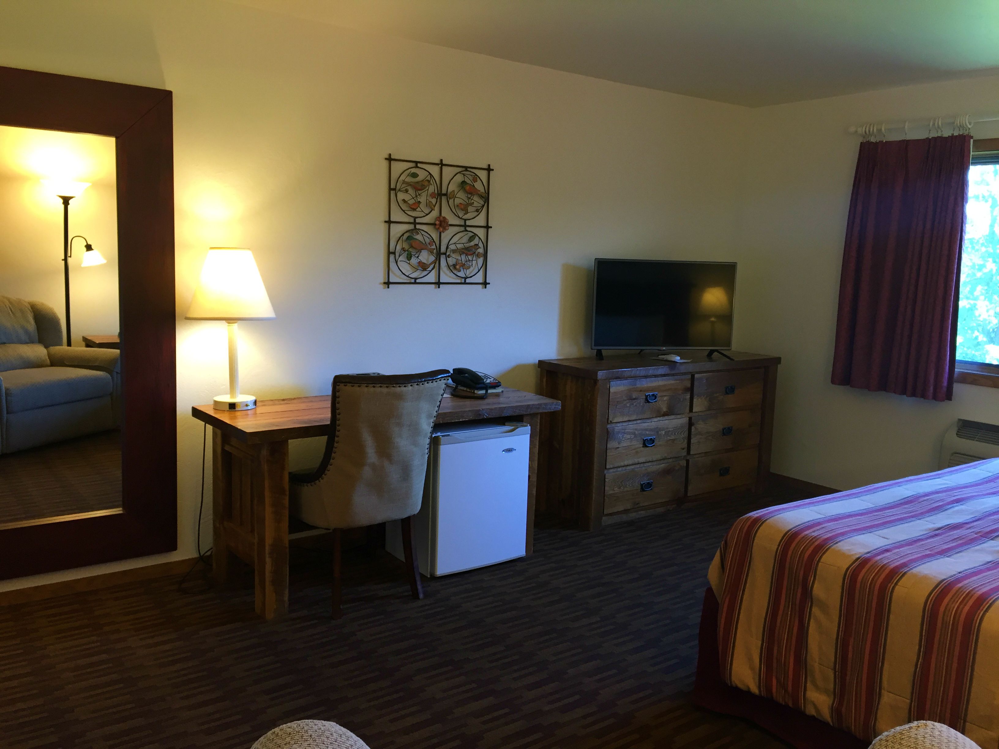 Une chambre d’hôtel comprenant un lit, un bureau avec une chaise, une télévision, un petit réfrigérateur, une lampe et un élément décoratif mural.
