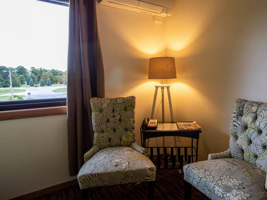 Una habitación con dos sillones tapizados, una lámpara de pie y un ventanal con vistas a una carretera y árboles.