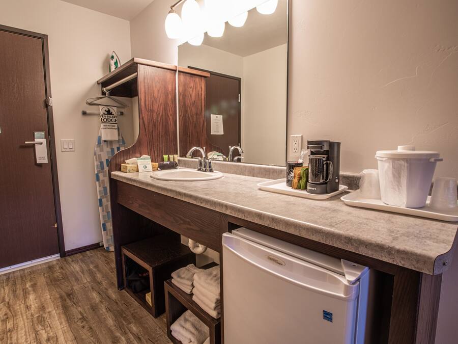 A hotel bathroom with a sink, mirror, and coffee maker. There is a towel rack and a white refrigerator.