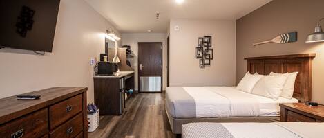 Habitación de hotel con una cama grande, una cama más pequeña, un escritorio, una cómoda, un televisor y un baño visible a través de una puerta abierta.