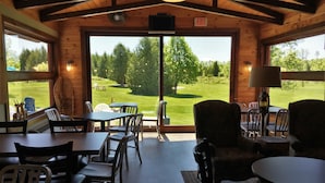 Interior acogedor de una cabaña con paredes de madera y ventanas amplias que ofrecen vistas a un patio cubierto de césped y árboles.