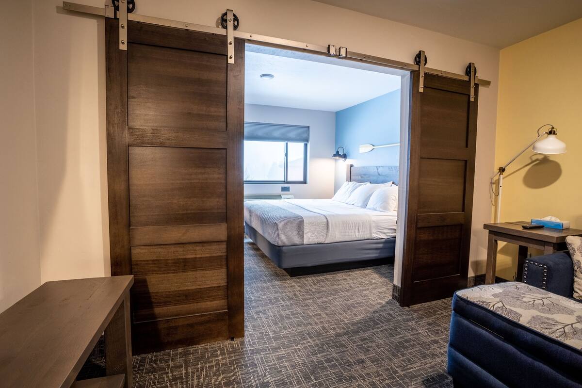 A hotel room with a sliding door leading to a bedroom, a desk with a lamp, and a patterned carpet.