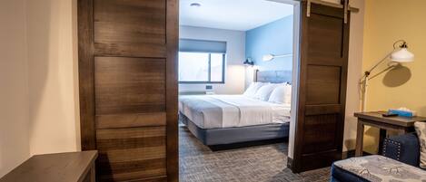 A hotel room with a sliding door leading to a bedroom, a desk with a lamp, and a patterned carpet.