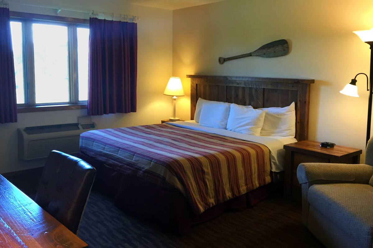 A hotel room with a bed, a wooden headboard, a nightstand with a lamp, a chair, a window with curtains, and a wall-mounted paddle.