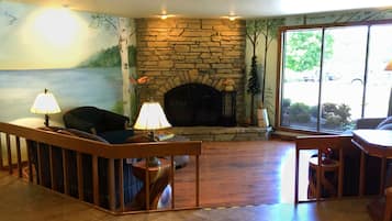 A living room with a stone fireplace, wooden flooring, and a view of the outdoors through large windows.