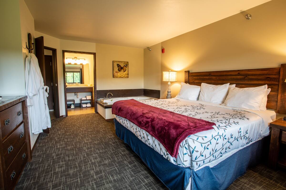A hotel room with a large bed, a wooden headboard, a nightstand, a bedside lamp, a bathroom with a mirror, and a robe hanging on a hook.