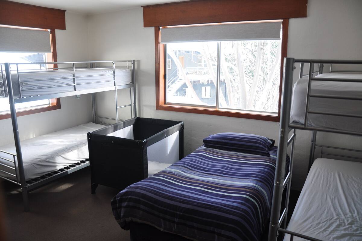 A dormitory room with bunk beds, a small desk, and a window with a view of buildings and trees.