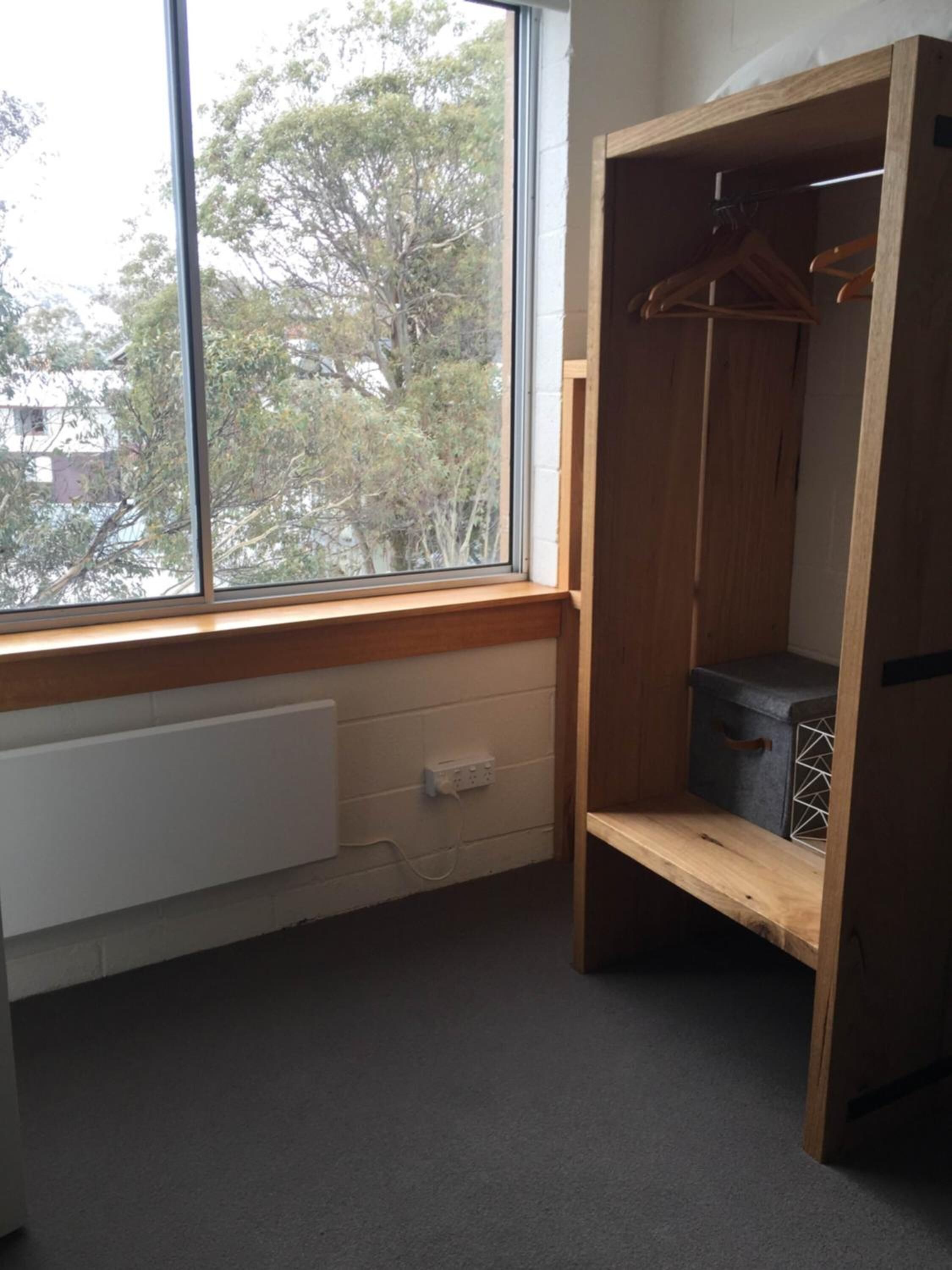 A room with a large window, a wooden wardrobe, a radiator, and a grey storage box.