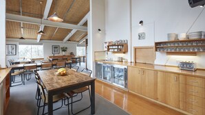 A spacious kitchen with wooden cabinets, a dining area with tables and chairs, and a large window providing natural light. - Ripparoo Ski Lodge (Falls Creek)