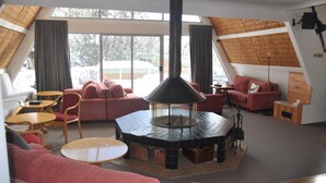 A cozy living room with a central fireplace, red sofas, a coffee table, and a large window offering a view of the outside. - Ripparoo Ski Lodge (Falls Creek)