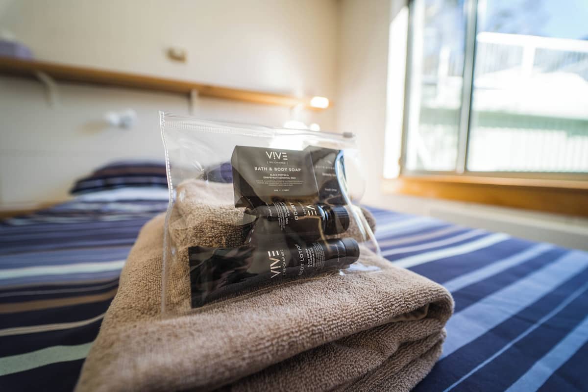 A neatly folded towel on a bed with a striped bedspread. In front of the towel, there is a clear plastic bag containing toiletries including a bottle of bath and body soap.