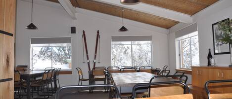 A spacious dining area with a large wooden table and chairs, high ceilings with exposed beams, and large windows providing natural light.