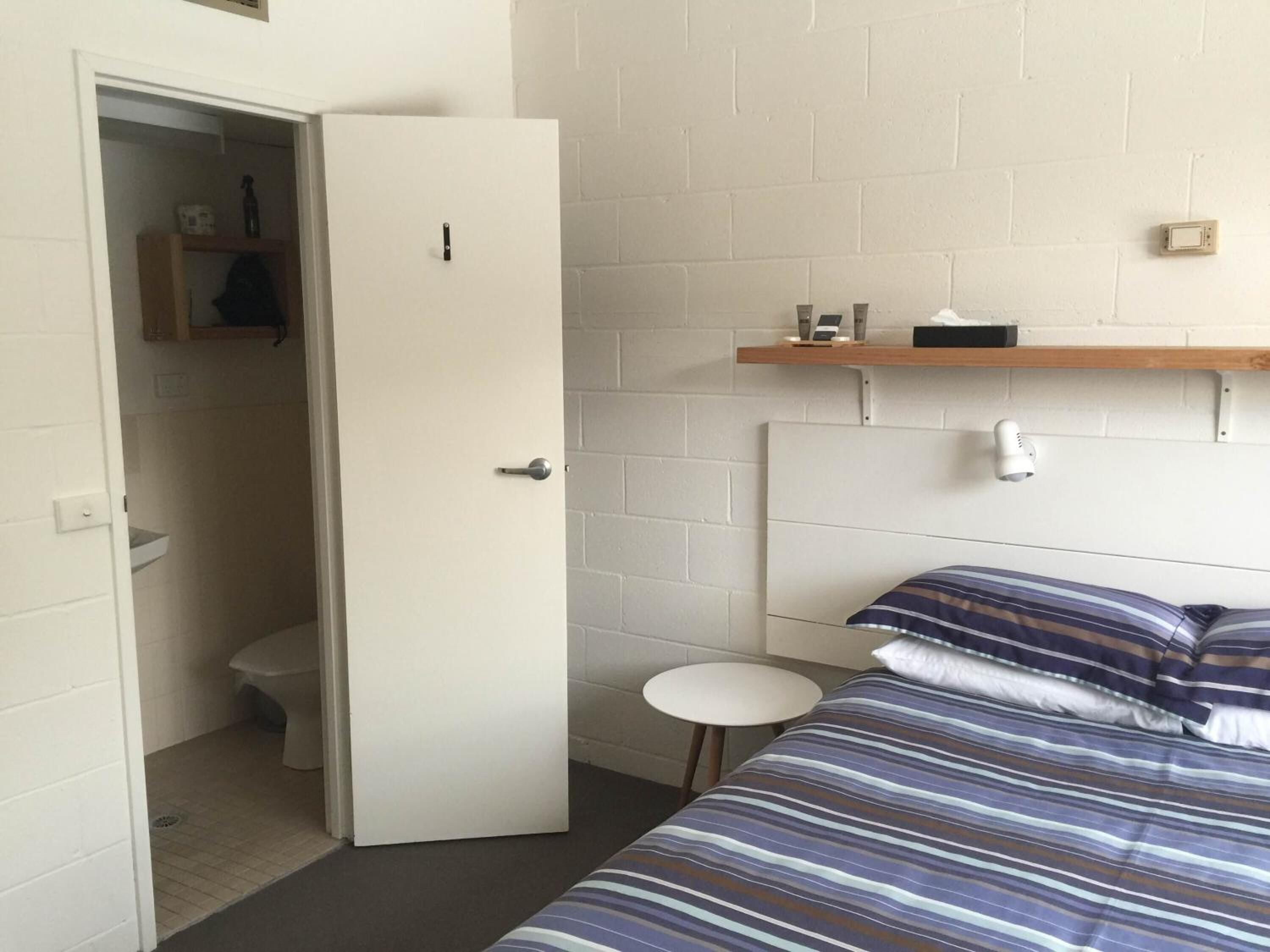 A small, clean room with a bed, a bedside table, a small stool, a shelf with toiletries, and a door leading to a bathroom.