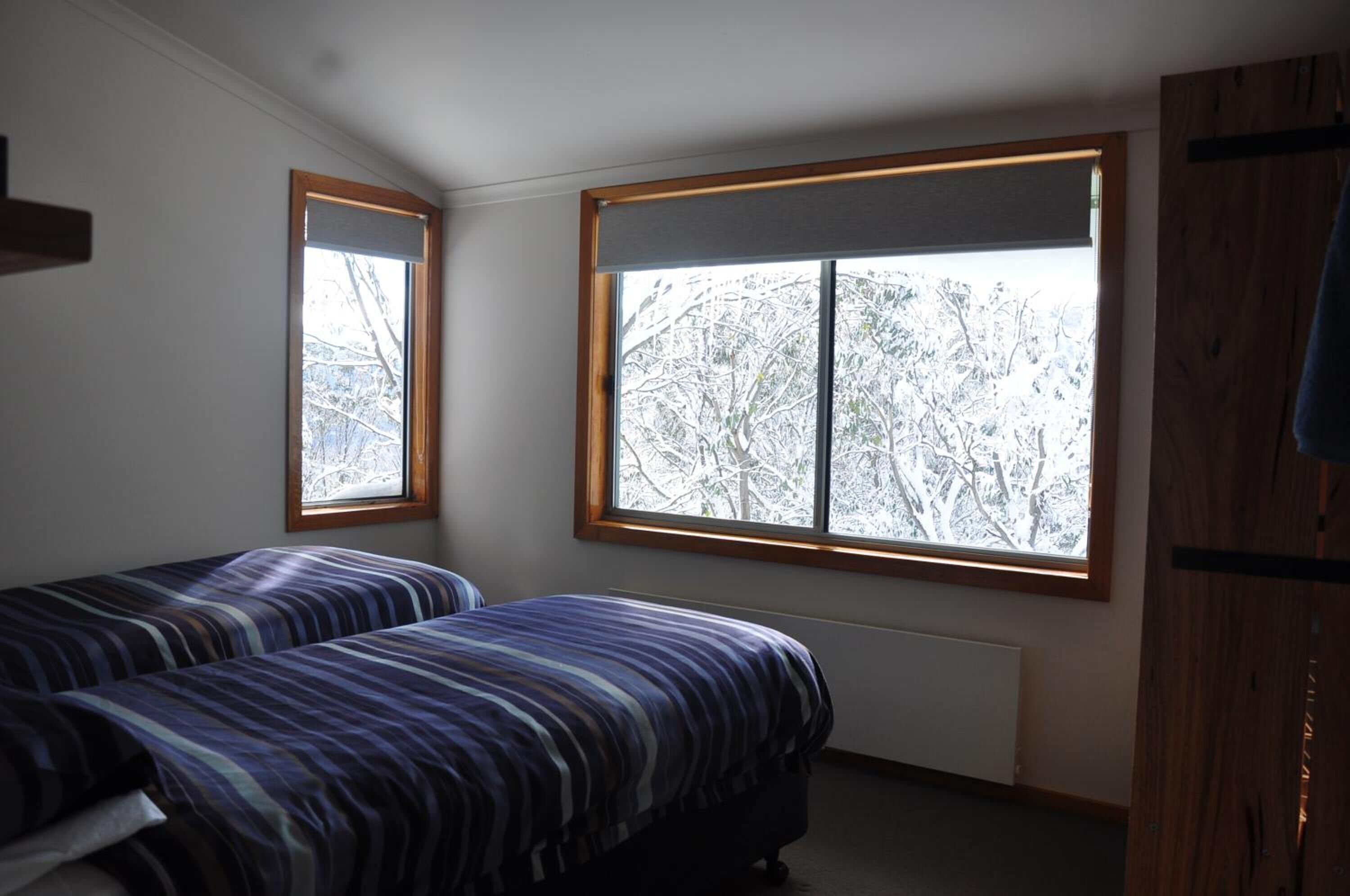 A bedroom with two beds, large windows with wooden frames, and a view of snow-covered trees.