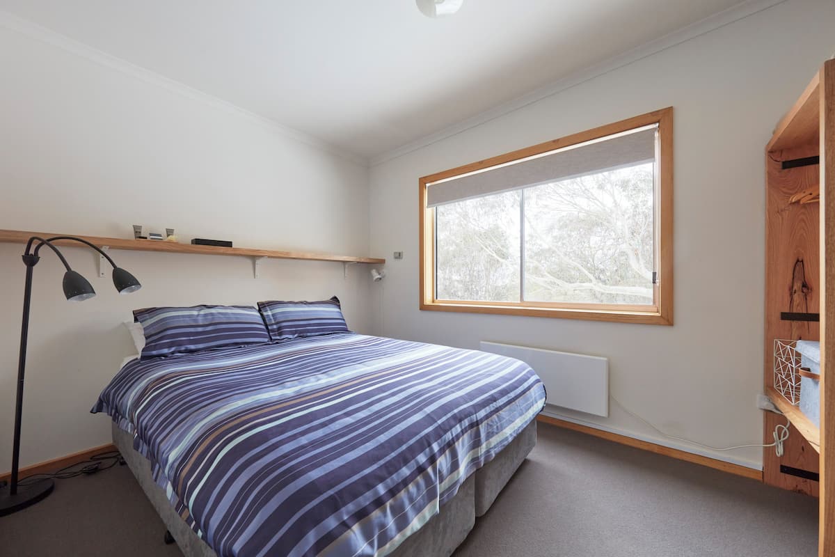 A bedroom with a bed, a window, a radiator, a shelf, and a desk.