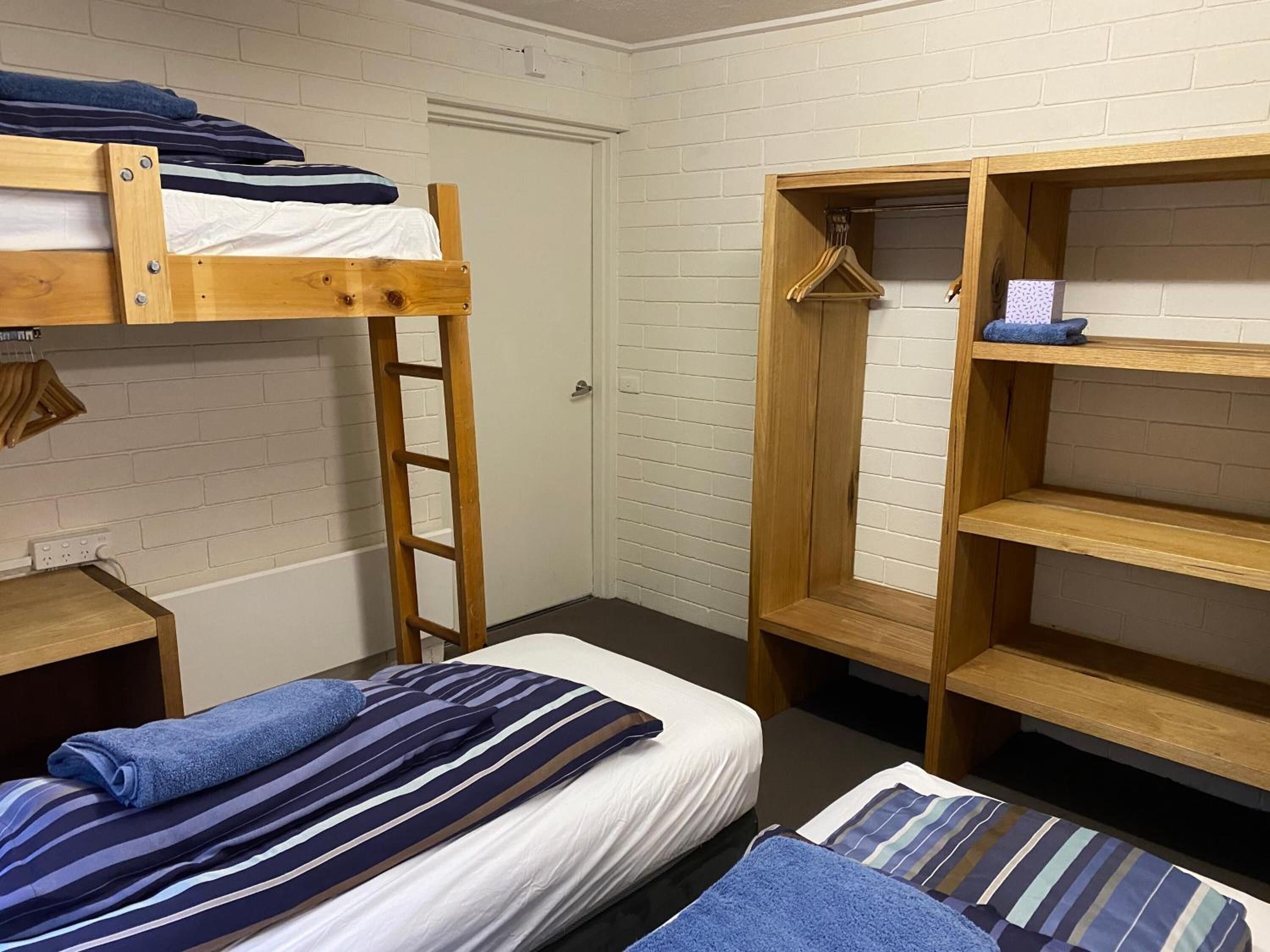 A dormitory room with bunk beds, a wooden ladder, a closet with hanging clothes, and a small desk.