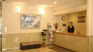 A reception area with a receptionist, a reception desk, a bench, a potted plant, a wall clock, and a framed artwork. - Toyooka Park Hotel (Toyooka)