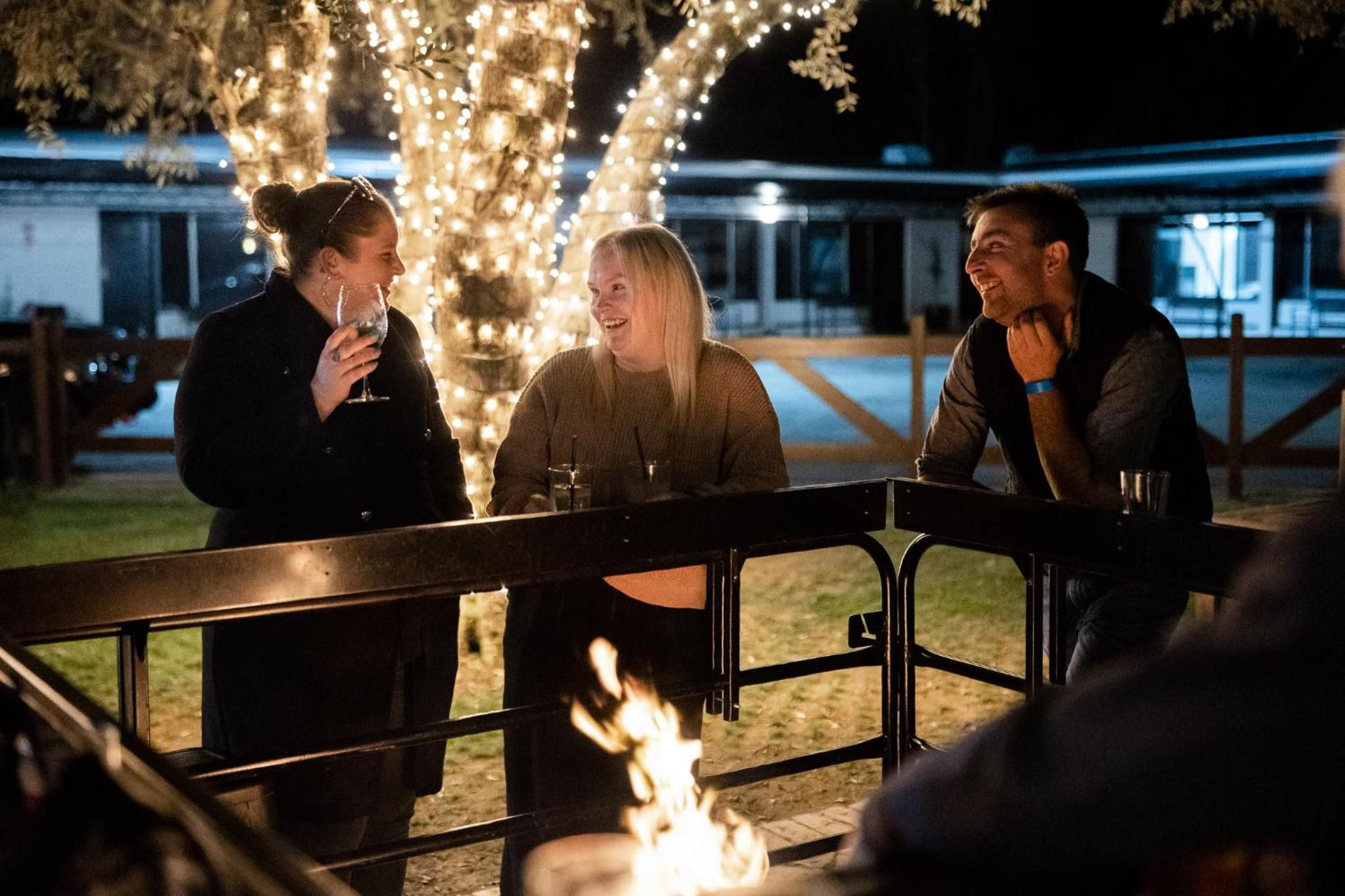 Three people seated at an outdoor table with a fire in the foreground, surrounded by trees and a fence.