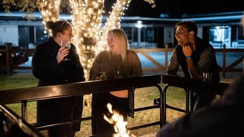 Three people seated at an outdoor table with a fire in the foreground, surrounded by trees and a fence.