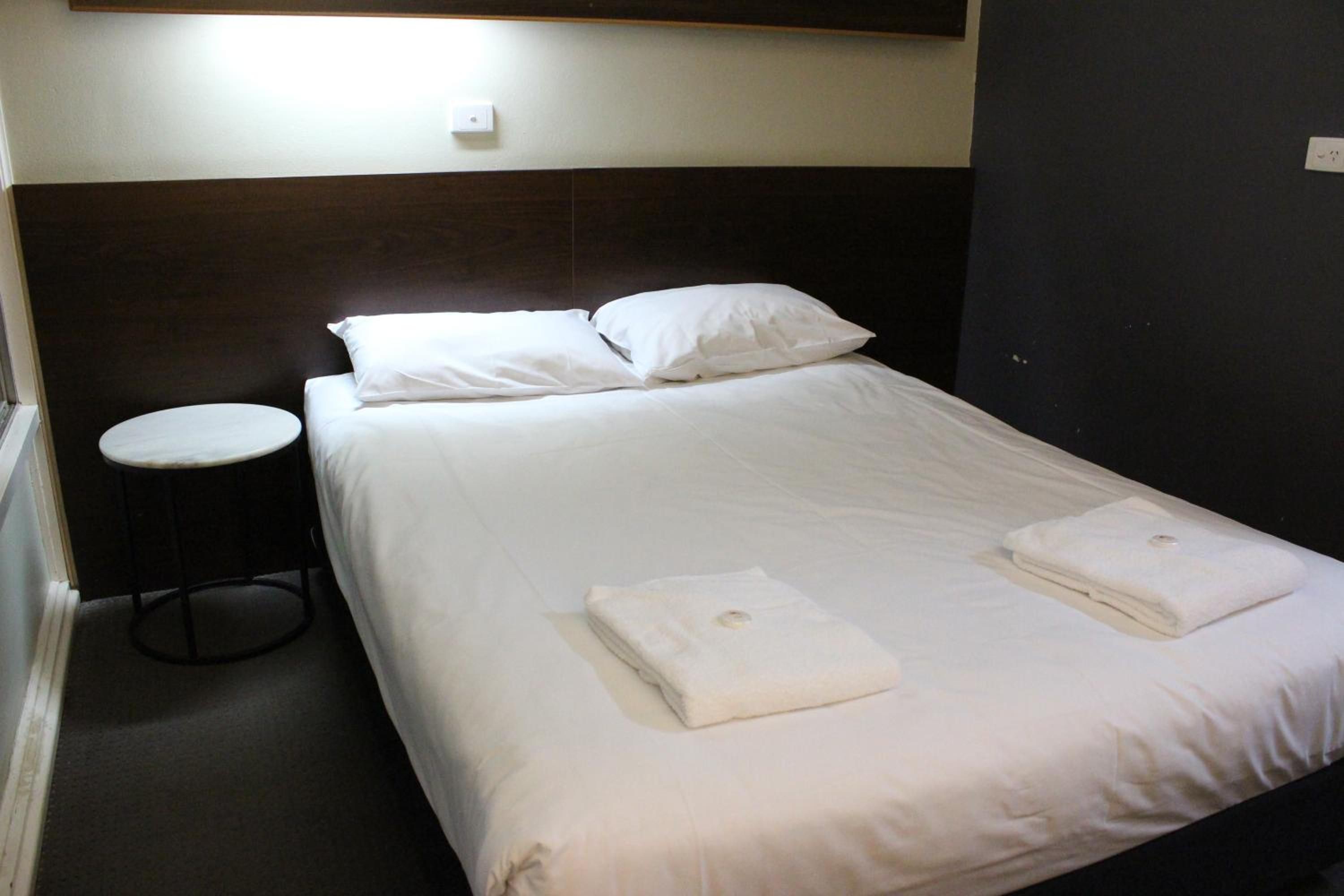 A single bed with white linens and two folded towels, a round stool, and a wall-mounted light fixture.