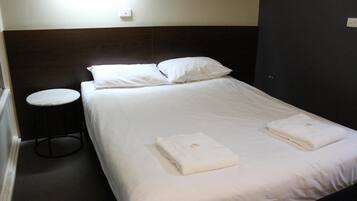 A single bed with white linens and two folded towels, a round stool, and a wall-mounted light fixture.