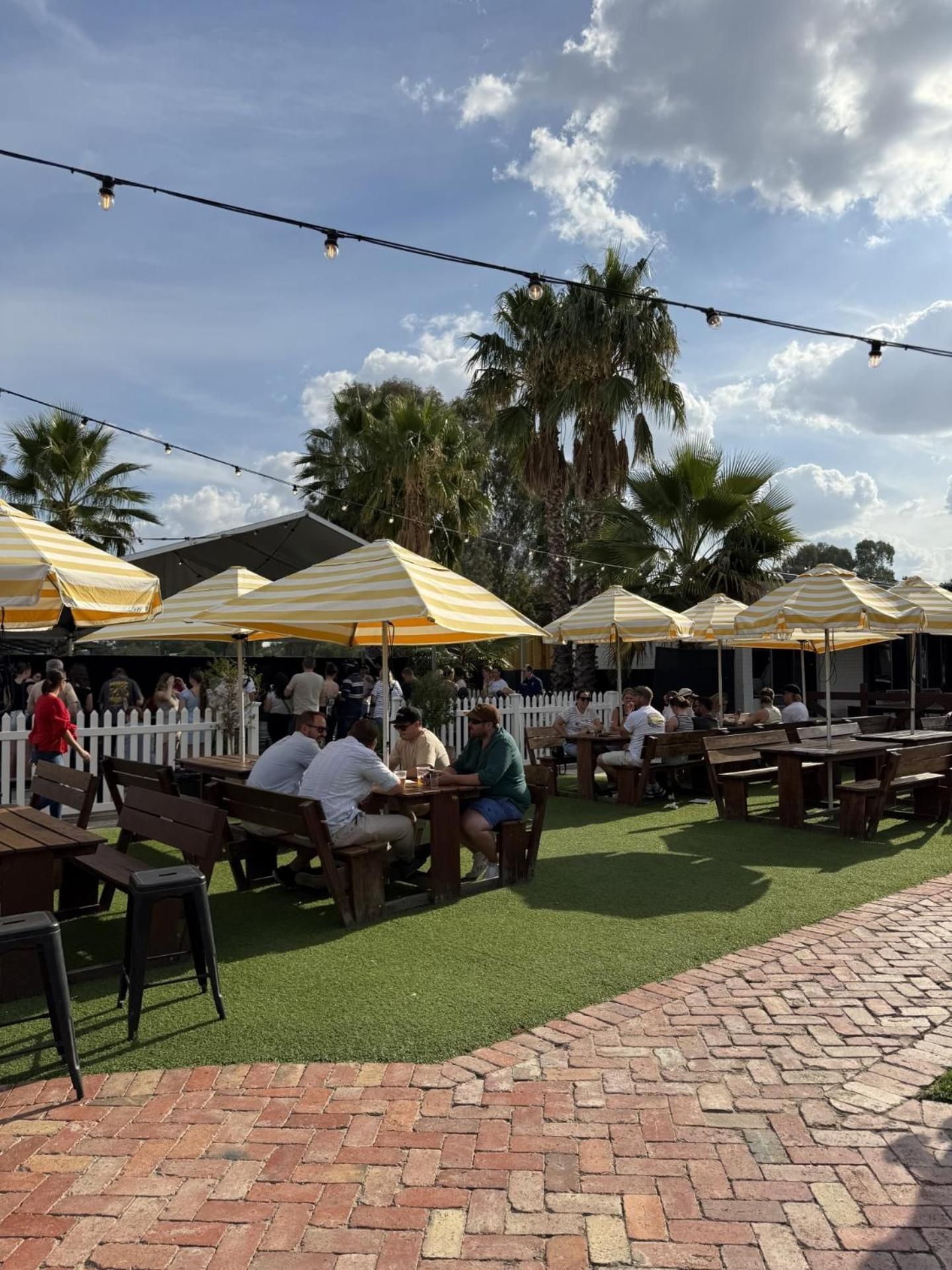 An outdoor seating area with tables and chairs, surrounded by palm trees and a white picket fence.