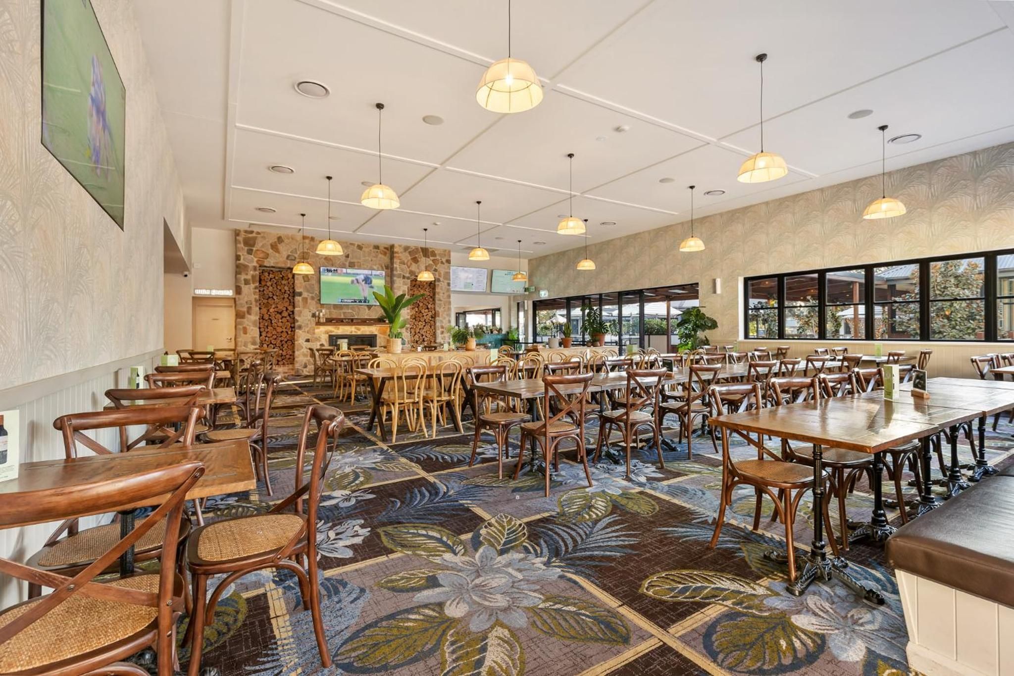 A spacious dining area with multiple tables and chairs, a stone wall with a fireplace, and large windows providing natural light.