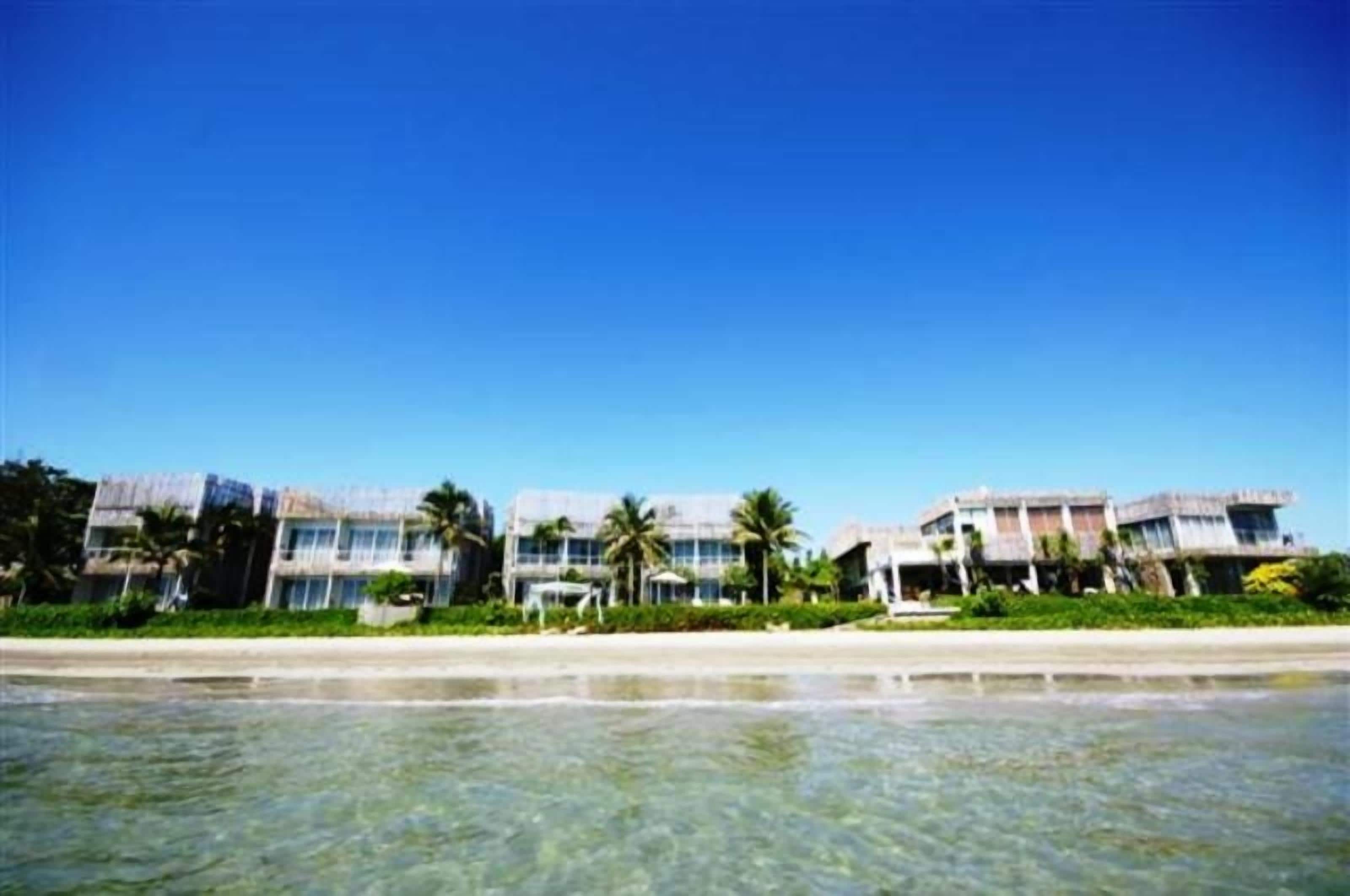 Un hotel frente a la playa con edificios de varios pisos, palmeras y una vista despejada del océano.