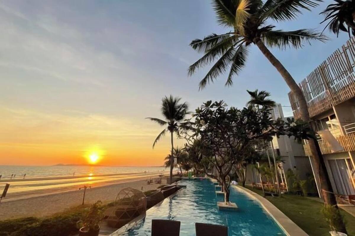 Piscina con sillas de descanso y palmeras, con vista a una playa al atardecer.