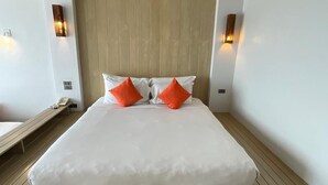 Una cama grande con ropa blanca y dos almohadas anaranjadas, suelo de madera y dos lámparas fijadas a la pared.
