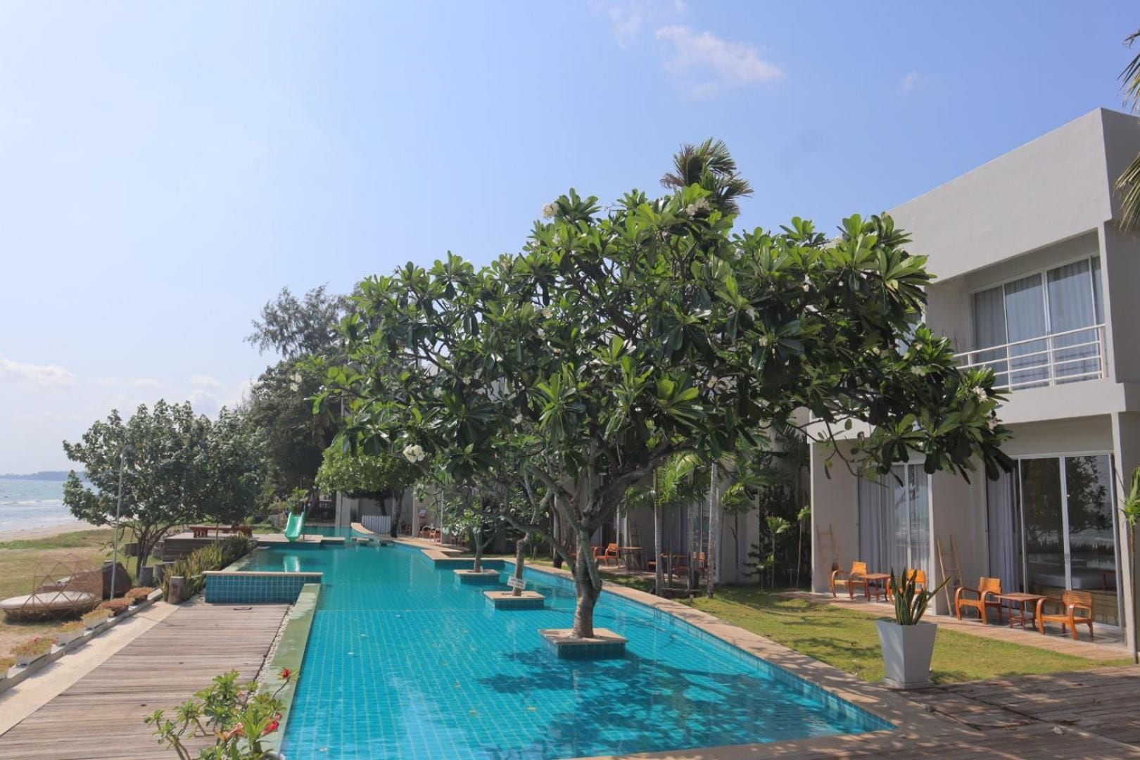 A swimming pool with a wooden deck, surrounded by greenery and a building with large windows.