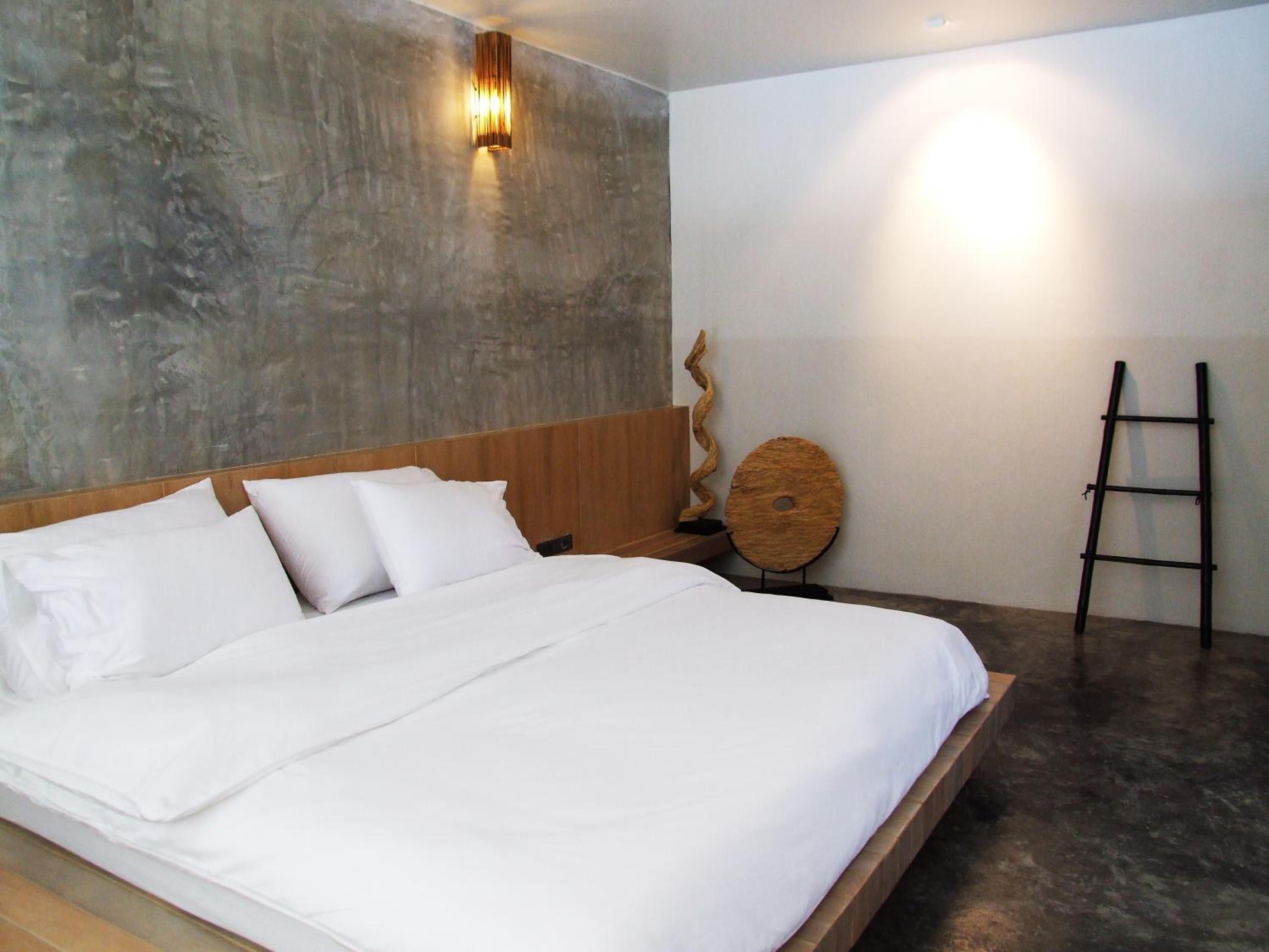 A bedroom with a large bed, a wooden headboard, a concrete wall, a round wooden object, and a black ladder.