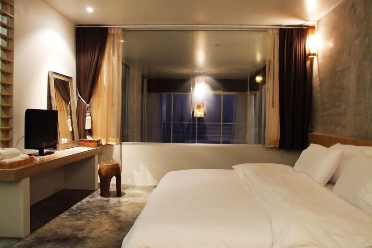 A hotel room with a bed, a desk with a computer, a chair, a mirror, and a window with curtains.