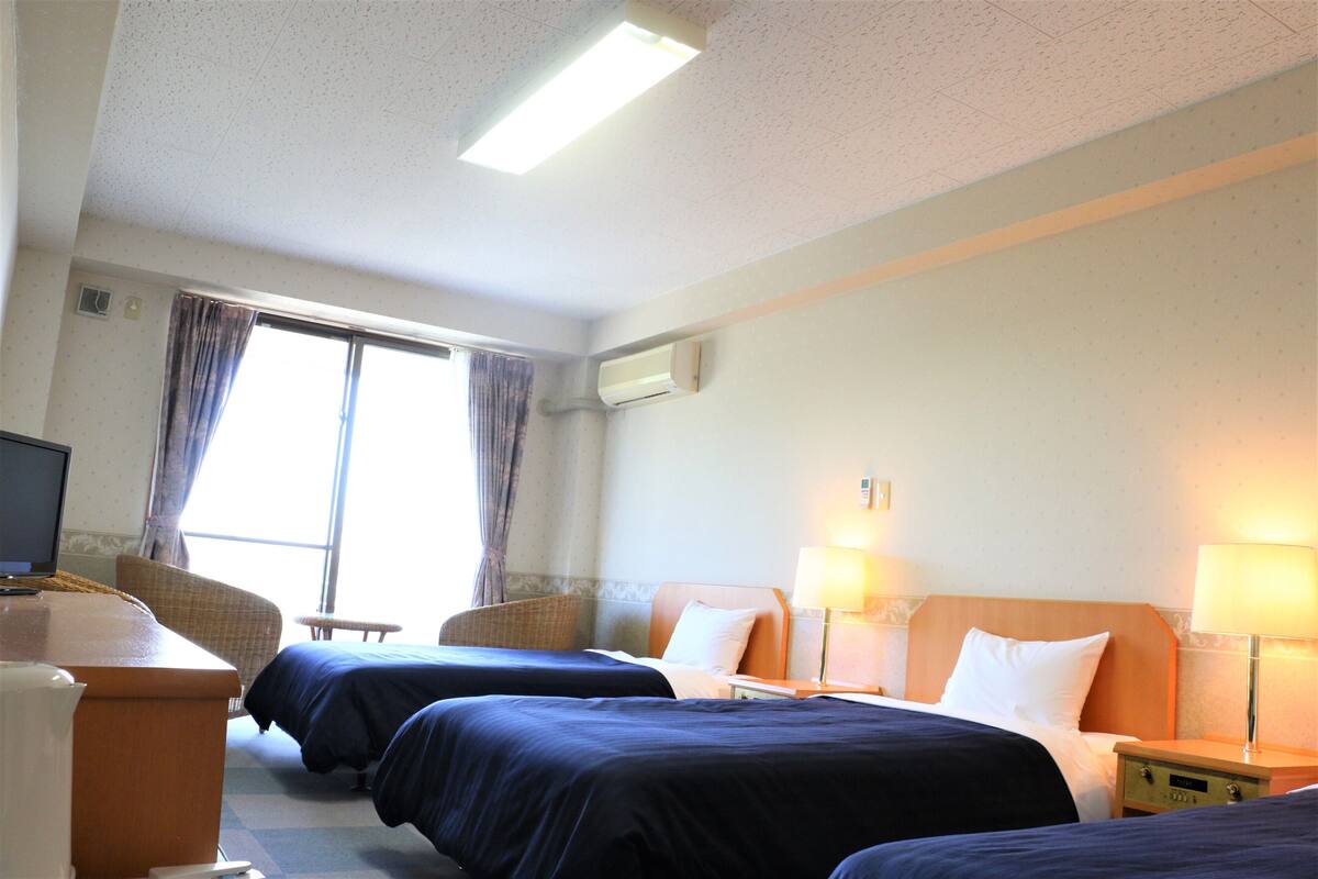 Una habitación de hotel con dos camas, un escritorio con computadora, una silla y una ventana con cortinas.