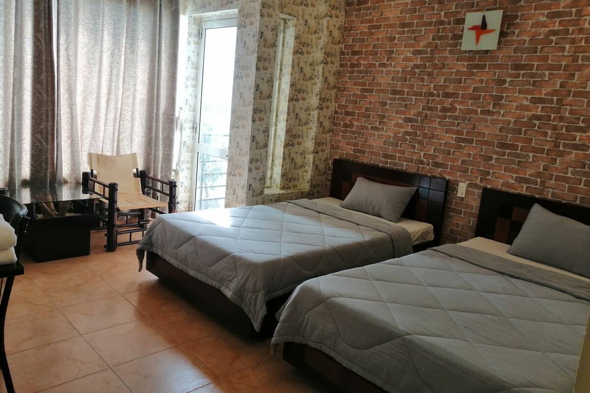 Una habitación con dos camas, una silla, una mesa y un ventanal con cortinas.