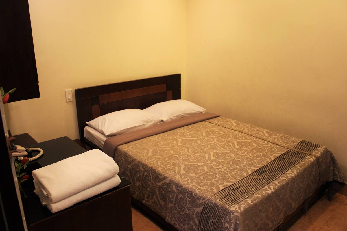 Habitación de hotel con cama, mesita de noche con teléfono y lámpara, e enchufe en la pared.
