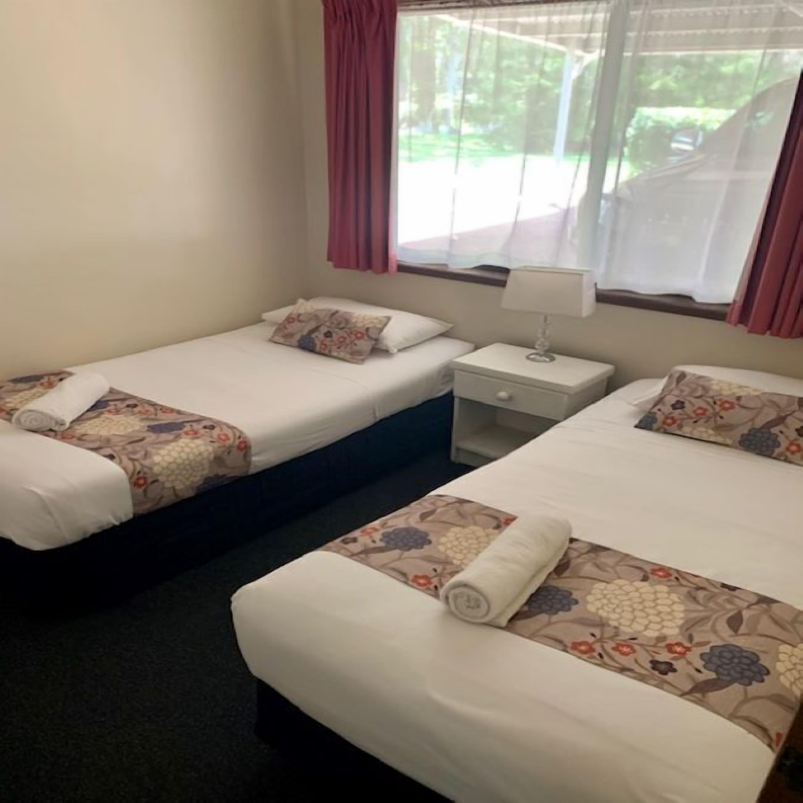 A hotel room with two beds, each with floral-patterned bedding and pillows, a bedside table with a lamp, and a window with curtains.