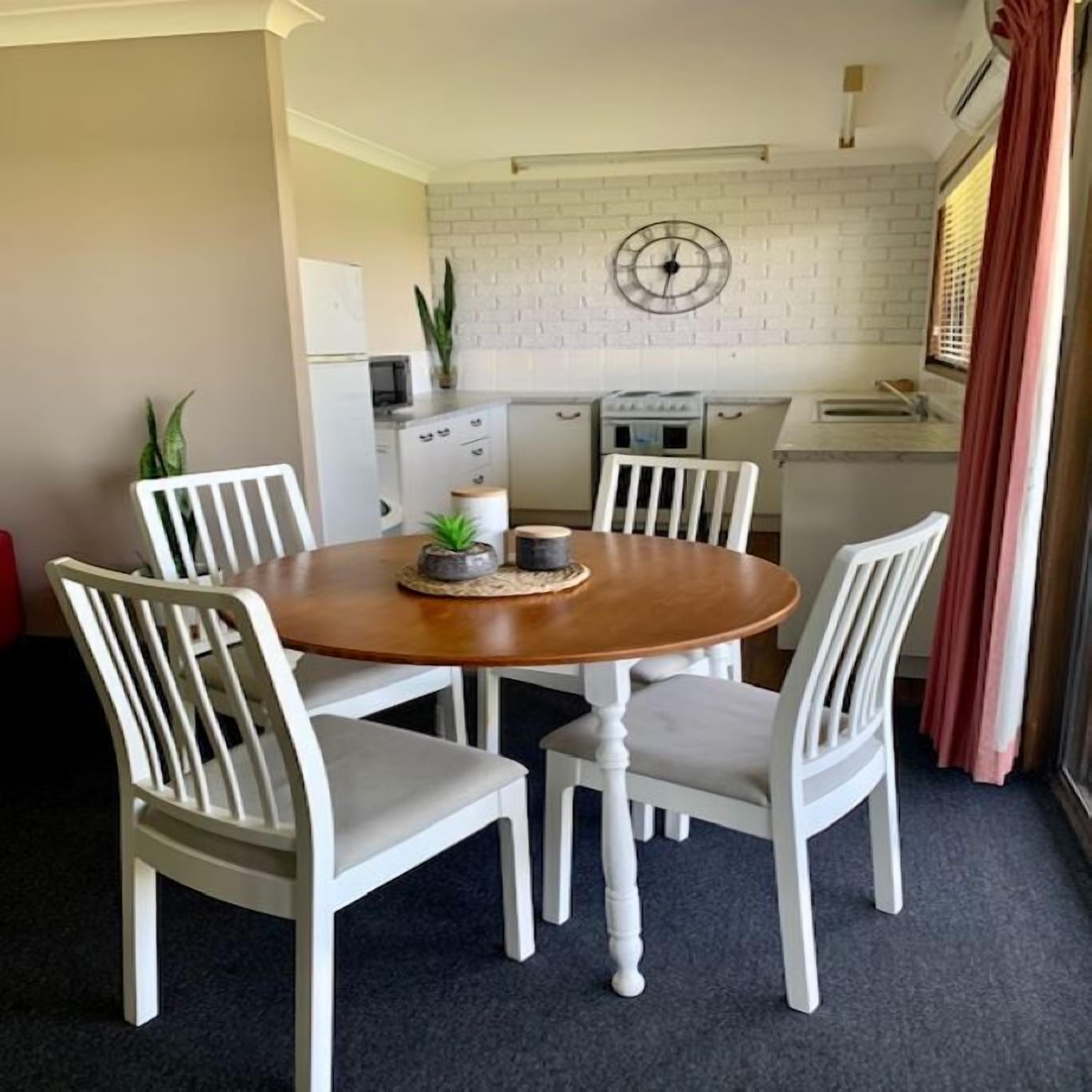 A dining area with a round wooden table surrounded by white chairs. A clock is mounted on the wall above the table.