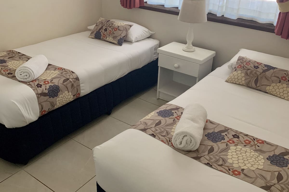 A hotel room with two single beds, each with a floral-patterned bedspread and a rolled towel. There is a small bedside table with a lamp, a window with curtains, and a view of greenery outside.