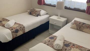 Habitació d'hotel amb dos llits individuals, cadascun amb una colcha de flors i una tovallola enrotllada. Hi ha una petita taula de nit amb una làmpada, una finestra amb cortines i vistes a la vegetació.