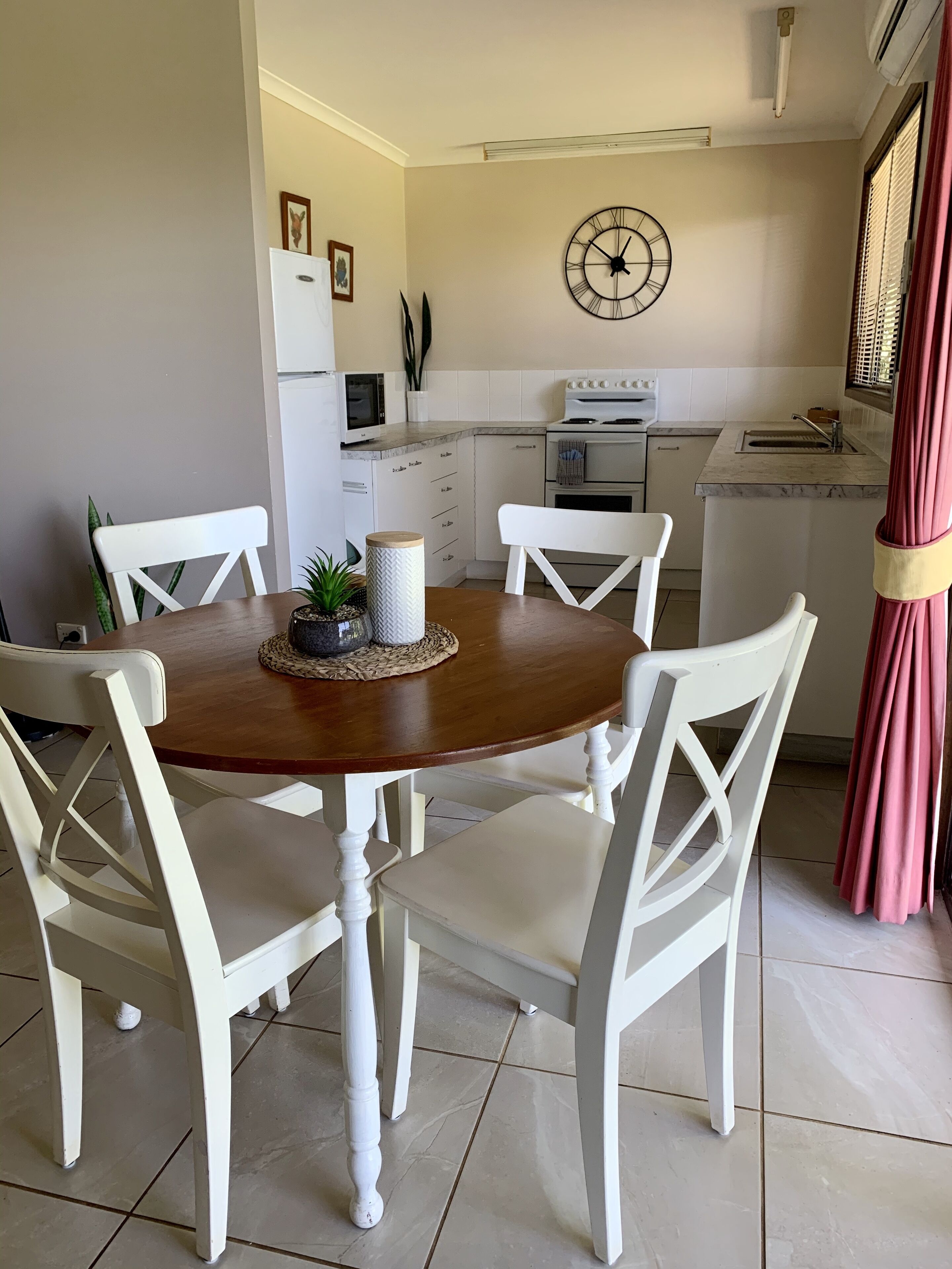 Een eetgedeelte met een houten tafel en witte stoelen, een keuken op de achtergrond en een klok aan de muur.