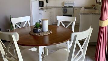 Een eetgedeelte met een houten tafel en witte stoelen, een keuken op de achtergrond en een klok aan de muur.