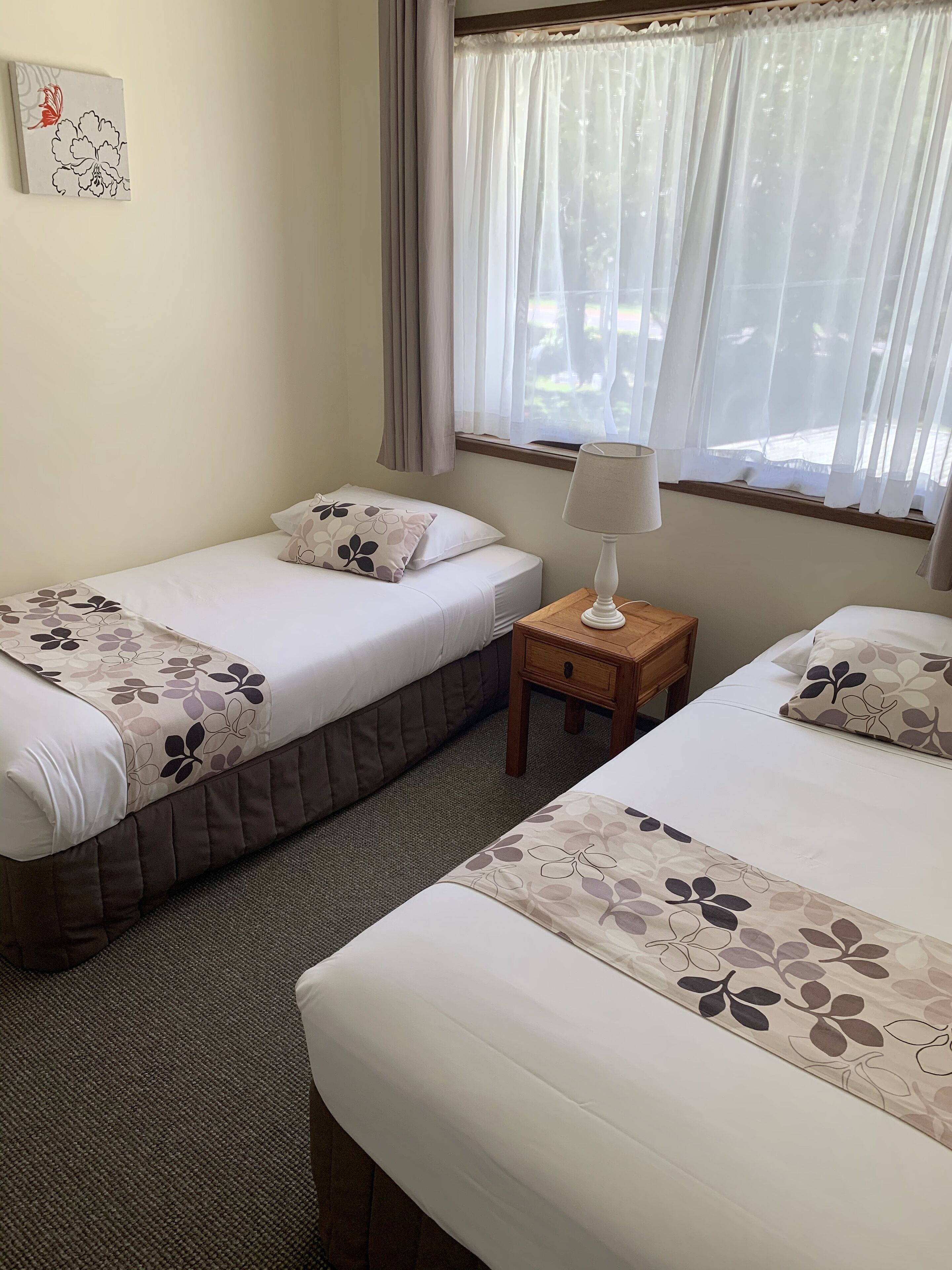 A hotel room with two single beds, a bedside table with a lamp, a window with sheer curtains, and a wall-mounted artwork.