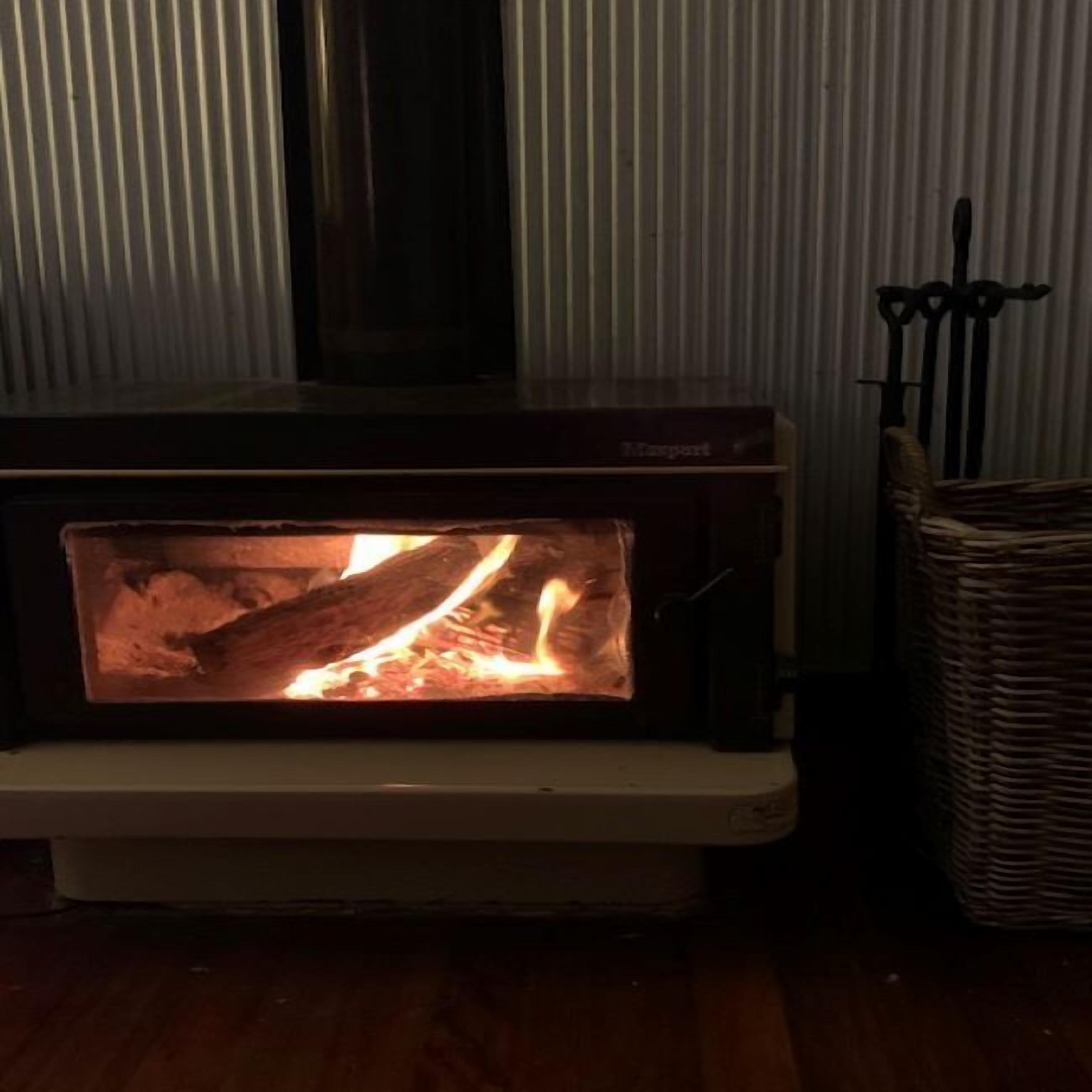 A wood-burning stove with visible logs and flames, a wicker basket to the right, and a metal railing behind.