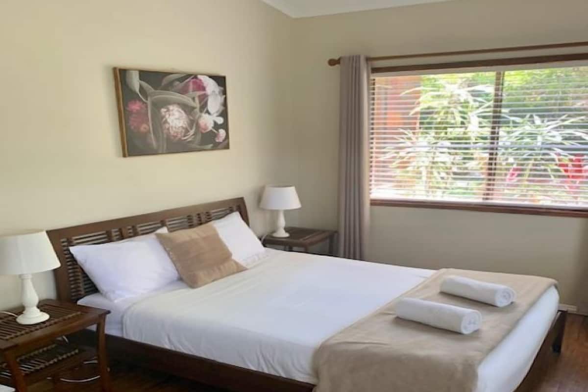 A neatly made bed with white linens, a beige throw blanket, and two rolled towels on the side. There is a wooden headboard, a nightstand with a lamp, and a painting on the wall.