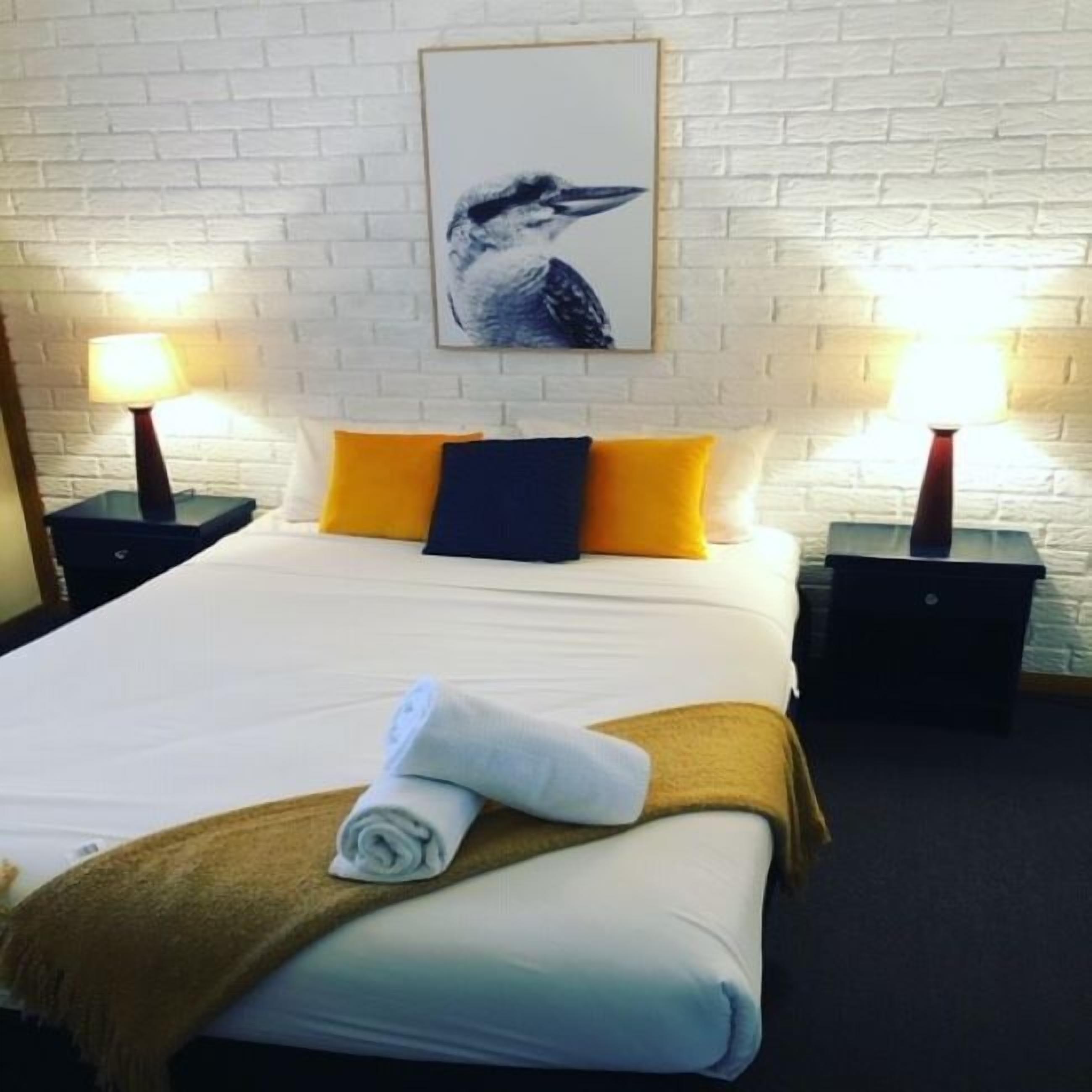 Habitación de hotel con pared de ladrillo blanco, cama con ropa blanca, dos almohadas amarillas, una almohada azul oscuro y una toalla enrollada sobre la cama. Hay dos mesitas de noche con lámparas, y un cuadro de un pájaro en la pared.