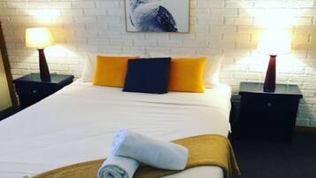 A hotel room with a white brick wall, a bed with white bedding, two yellow pillows, a dark blue pillow, and a rolled towel on the bed. There are two bedside tables with lamps, and a framed picture of a bird on the wall.